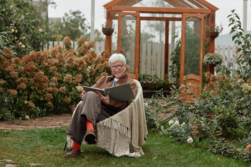 Senior Caucasian man sitting outdoors in village garden holding large photo album, smiling while...