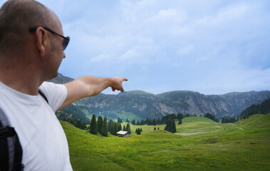 Man in sunglasses gestures towards a scenic landscape with rolling green hills, trees, and a...
