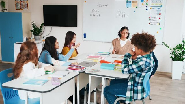 A female teacher sits at a desk with a group of five students sitting behind it, they are talking