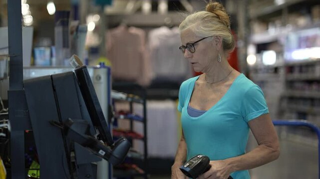 A confused, frustrated unhappy, mature woman stands at a self checkout station in a hardware store holding a hand scanner.