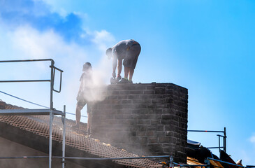 Two construction workers demolish a heavy chimney using a cutting machine, raising dense dust...