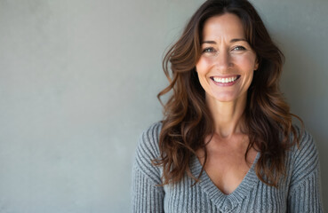Mature woman smiling. Middle-aged female with long brown hair poses in grey sweater against neutral background. Happy face, positive emotions, good mood, beauty and lifestyle concept.