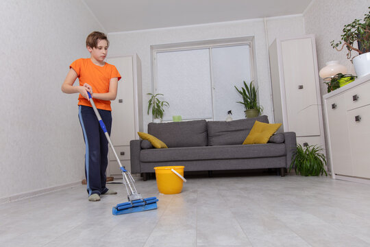Young boy cleans living room floor with mop and bucket in bright, modern home during daytime - Powered by Adobe