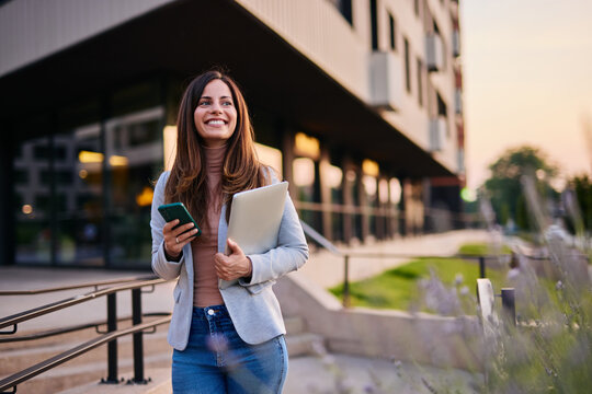Smiling Woman Holding Laptop and Phone Outside Modern Building During Sunset