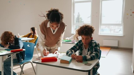 A female teacher stands at a desk with a schoolboy sitting behind it, they are writing in notebooks - Powered by Adobe