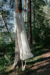 white dress is gracefully hung from a tree branch surrounded by lush greenery. Sunlight filters through the forest, creating a tranquil atmosphere in the afternoon