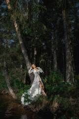 woman stands elegantly in a white dress beside a river, surrounded by tall trees. Sunlight filters...
