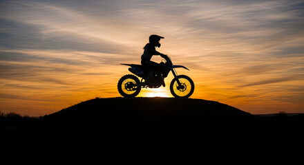 Obraz premium Silhouette of a motocross rider on a dirt bike atop a hill against a vibrant sunset sky.