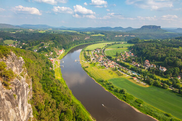 Scenic panoramic view from the Bastei cliffs over the Elbe River valley with sandstone rocks, lush forests, rural village rooftops, distant mountain ranges under a vibrant summer sky. Saxony, Germany