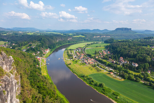 Scenic panoramic view from the Bastei cliffs over the Elbe River valley with sandstone rocks, lush forests, rural village rooftops, distant mountain ranges under a vibrant summer sky. Saxony, Germany