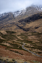 A solitary figure traverses the rugged, winding path through the majestic and mist-shrouded peaks of the Scottish Highlands, possibly in the iconic Glen Coe, where ancient landscapes meet dramatic ski