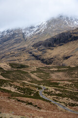 A solitary figure traverses the rugged, winding path through the majestic and mist-shrouded peaks of the Scottish Highlands, possibly in the iconic Glen Coe, where ancient landscapes meet dramatic ski