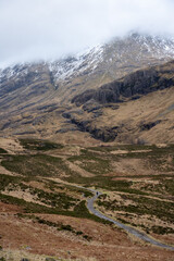 A solitary figure traverses the rugged, winding path through the majestic and mist-shrouded peaks of the Scottish Highlands, possibly in the iconic Glen Coe, where ancient landscapes meet dramatic ski