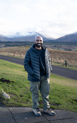A guy in a scenic view of a road leading into a vast, rugged landscape with snow-capped mountains under a cloudy sky, likely in the Scottish Highlands