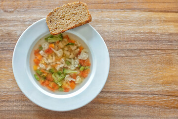 Top view of clear vegetable soup with a slice sourdough bread on white plate on wooden surface