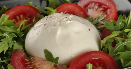 Burrata cheese with tomatoes and arugula is sprinkled with salt and dried chili pepper. Close-up footage on the rotating table.