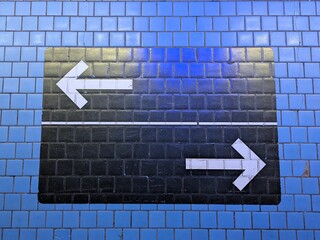 Urban signage painted on a blue tiled wall, featuring two directional arrows pointing left and...