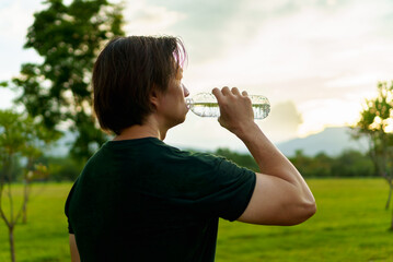 Athlete drinking water from a bottle after running in park, outdoor workout with light of sunset on freshy green of nature  background. Thirsty runner and bottle for hydration, electrolytes.