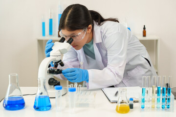Asian teenage girl dressed in white lab coat and protective goggles studies a specimen under...