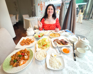 Woman enjoying traditional chinese food at restaurant, A european girl in a red dress sitting at a table with a variety of Asian dishes, smiling and relaxed.