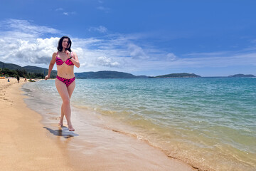 A young, happy, athletic woman in a bright pink swimsuit walks along a tropical beach, a girl runs along a sandy beach against the backdrop of a spectacular sea landscape 