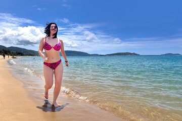 A young, happy, athletic woman in a bright pink swimsuit walks along a tropical beach, a girl runs along a sandy beach against the backdrop of a spectacular sea landscape 