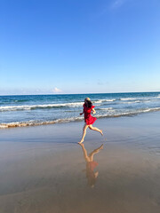 Woman in red dress running along the beach, A girl in a flowing bright dress joyfully jogging along the shore, reflected in the wet sand.