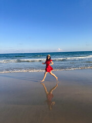 Woman in red dress running along the beach, A girl in a flowing bright dress joyfully jogging along the shore, reflected in the wet sand.