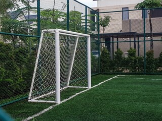 White football goal on green artificial turf field, surrounded by a fence with foliage and...