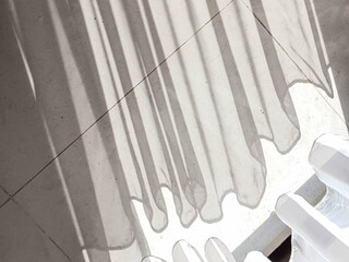 Sunlight streams through sheer curtains, casting long, dramatic shadows and playful wavy patterns on a textured light gray floor. A serene and minimalist abstract background.