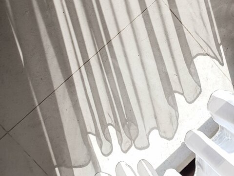 Sunlight streams through sheer curtains, casting long, dramatic shadows and playful wavy patterns on a textured light gray floor. A serene and minimalist abstract background. - Powered by Adobe