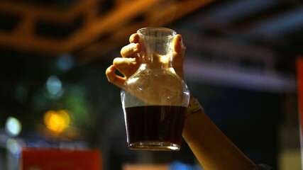 A hand holding a glass cup filled with cold coffee liquid, against a warm, hazy background with...