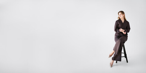 Smiling woman in brown medical uniform sitting on a stool on a wide white banner with space for text or design.