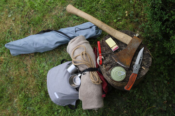 Hiking. Things and objects for tourism and survival in the forest or mountains. On the grass near the stump lie an axe, matches, canned food, rope, a tourist chair, a backpack and other things