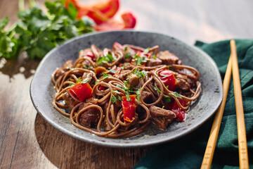 Wok with Soba noodles with turkey, mushrooms and sweet red bell pepper in plate on wooden table in the sun. Asian food, concept of street food.