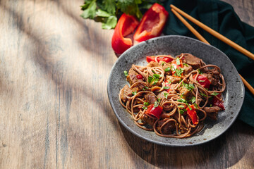 Wok with Soba noodles with turkey, mushrooms and sweet red bell pepper in plate on wooden table in the sun. Asian food, concept of street food.