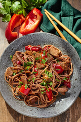 Wok with Soba noodles with turkey, mushrooms and sweet red bell pepper in plate on wooden table in the sun. Asian food, concept of street food. Top view. Close up.