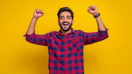 Fototapeta premium A young, handsome Indian man wearing a checkered shirt stands against an isolated yellow background, celebrating his victory with a winner's gesture