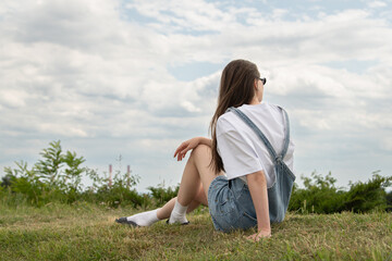 A young woman sits comfortably on a grassy hill, gazing into the distance. She wears casual summer attire and appears to be enjoying the serene atmosphere during a cloudy late afternoon