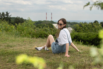 A young woman in casual attire sits on a grassy hill, enjoying the view of the distant landscape marked by smokestacks in the background under a cloudy sky