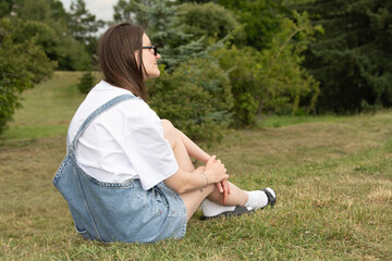 A woman sits on grass, wearing a simple white t-shirt and denim overalls. She appears relaxed as she enjoys the peaceful park surroundings on a mild day