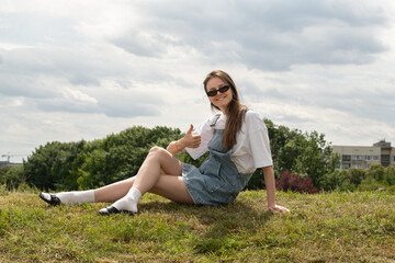 Bright day with scattered clouds, a young woman relaxes on grassy terrain. She wears denim...