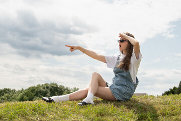 A young person wearing sunglasses and casual clothing is sitting on a grassy slope, pointing and looking toward the horizon. The sky is cloudy, creating a relaxed atmosphere