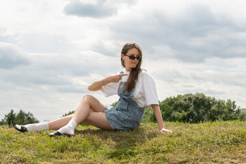 A young woman relaxes on grassy terrain wearing denim overalls and sunglasses. She strikes a playful pose under a partly cloudy sky, enjoying the outdoors on a warm day