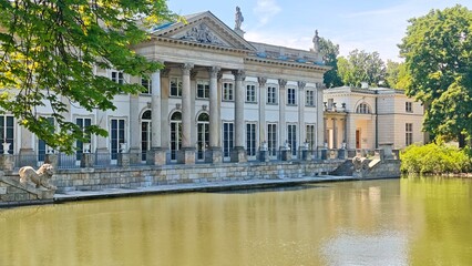 Classical palace with elegant columns and sculptures by a calm pond surrounded by green trees on a sunny day