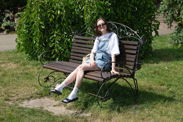 A woman relaxes on a metal bench in a vibrant garden, donning a casual outfit of a t-shirt and overalls. Sunlight filters through the trees, illuminating the scene