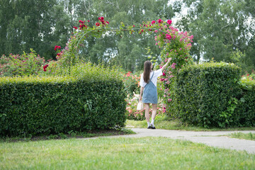 A young woman in a skirt walks through a beautiful rose garden filled with colorful blooms. She reaches out to touch some flowers while surrounded by greenery on a clear day