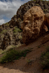 A large, weathered rock formation stands on a slope among rugged cliffs and sparse vegetation in...