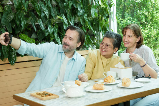 Asian senior woman and two Caucasian friends share laughter and smiles while taking a selfie at a cafe table, surrounded by pastries and tea, enjoying a happy retirement moment.