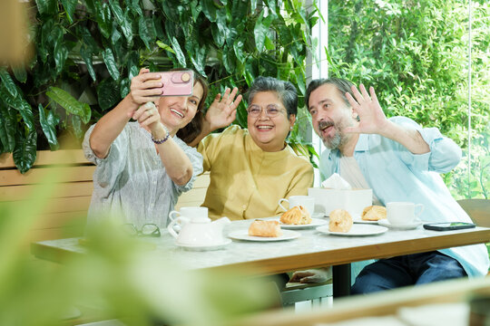 Asian senior woman and two Caucasian friends wave playfully while taking a selfie at a cafe table, laughing and enjoying pastries and tea in a joyful and relaxed retirement atmosphere.